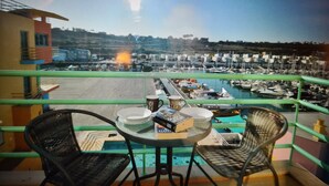 A balcony with two wicker chairs, a small round table, and a view of a marina with boats and a distant town.