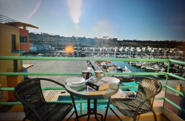 Un balcon avec deux fauteuils en osier, une petite table ronde et une vue sur une marina avec des bateaux et une ville située au loin.