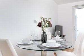En spiseplads med et glasbord, hvide stole, en vase med blomster og et fjernsyn i baggrunden.