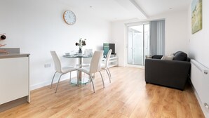Ein modernes Wohnzimmer mit einer Couch, einem Essbereich mit Glastisch und Stühlen sowie einem großen Fenster mit Vorhängen.