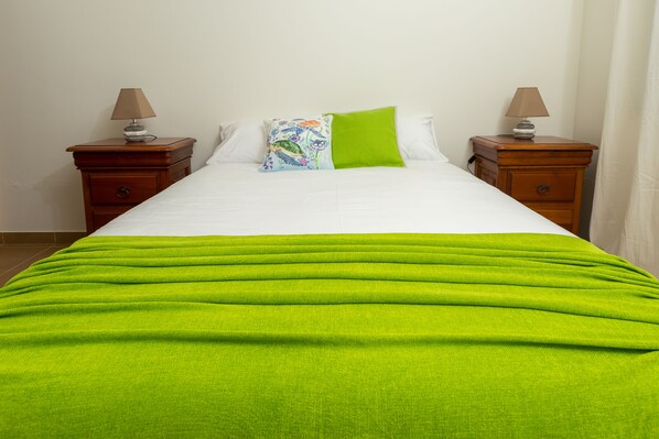 Un lit bien fait, recouvert d’une couverture verte, avec deux tables de chevet dotées de lampes et un coussin à motifs floraux.