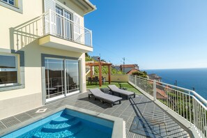 Un espace piscine avec des chaises longues, un patio fermé par des baies vitrées et une vue sur la mer.