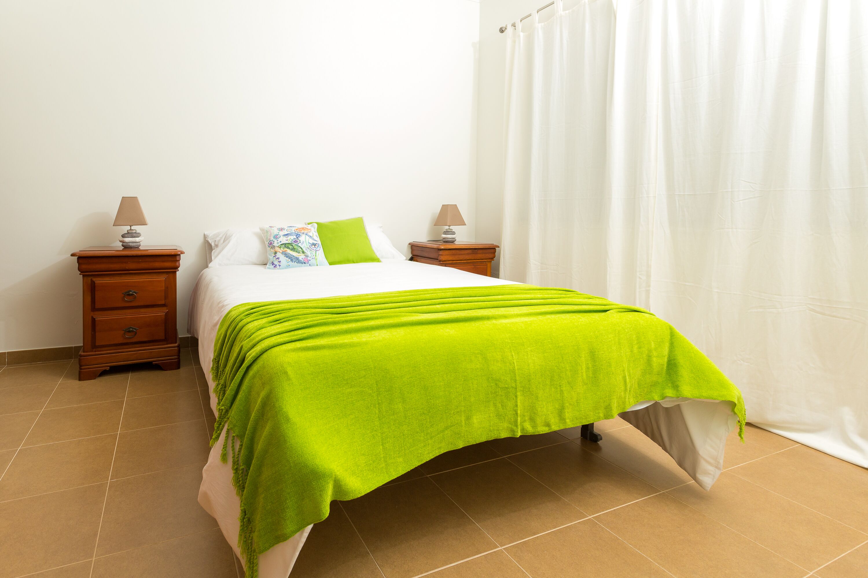 A neatly made bed with a bright green blanket, two bedside tables with lamps, and a white curtain.