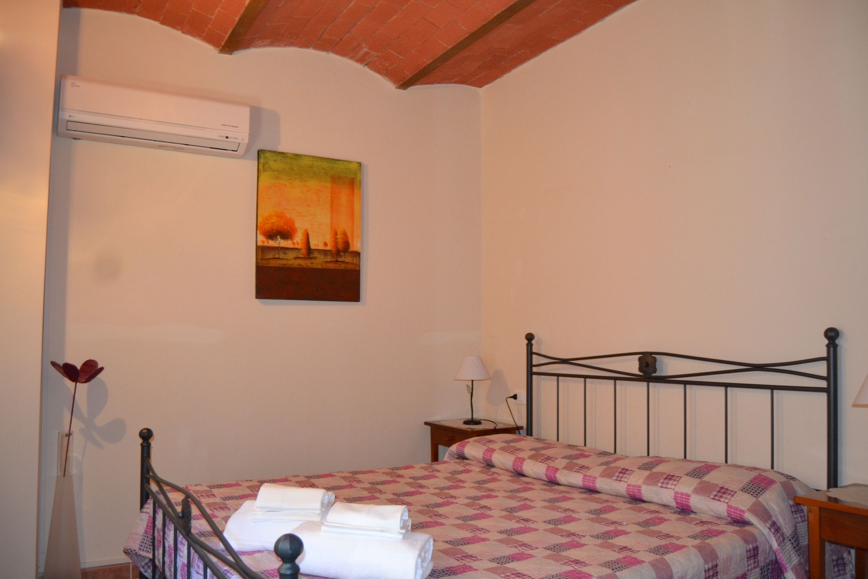 A bedroom with a bed, a nightstand, a lamp, an air conditioner, and a painting on the wall.