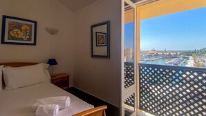 Habitación de hotel con cama, dos cuadros enmarcados, una mesita de noche y un balcón con vistas a un puerto deportivo.