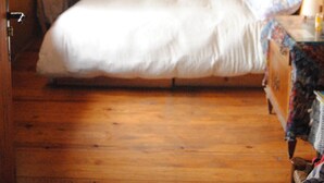 A bedroom with a wooden ceiling, a stone wall, a bed with white bedding, a window with white curtains, and a wooden dresser with a floral tablecloth. - Country house in Vall de Camprodon (Llanars)