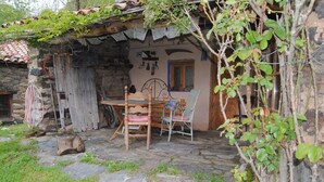 A rustic stone building with a tiled roof, a wooden table, two wicker chairs, and a small garden area with grass and a tree. - Country house in Vall de Camprodon (Llanars)