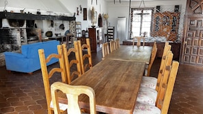 Un área de comedor con una larga mesa de madera y sillas a juego, un sofá azul y una cocina con pared de piedra y utensilios colgantes.
