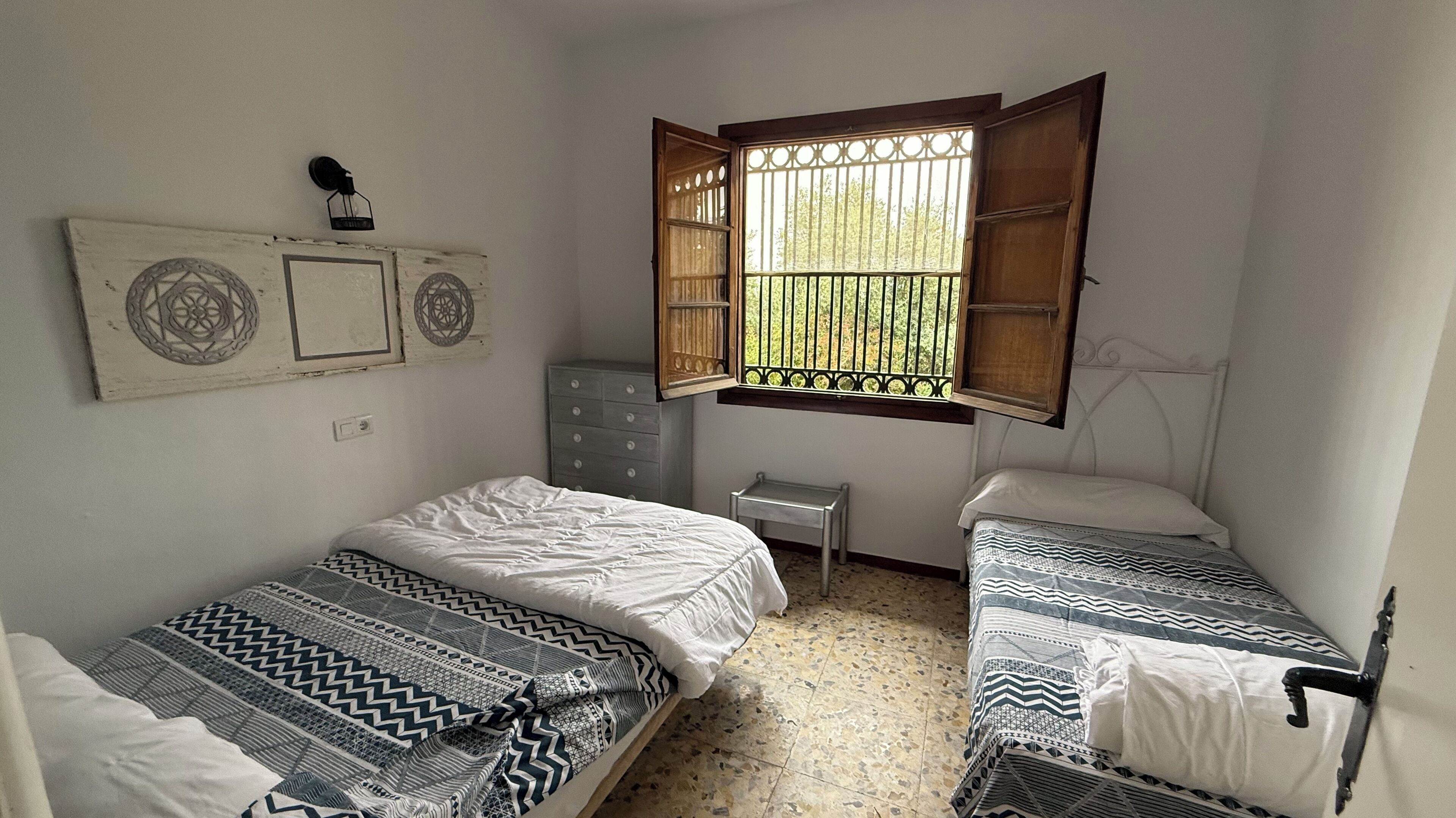 Una habitación pequeña y sencilla con dos camas, una mesita y una ventana con barrotes.