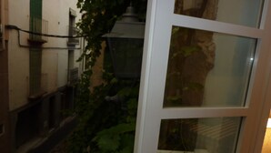 Un callejón estrecho con una puerta blanca y una ventana, parcialmente oculto por follaje verde.