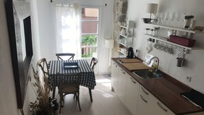 Cocina compacta con mesa, sillas y un lavamanos. Hay estantes con utensilios de cocina y una ventana con vistas a la vegetación.