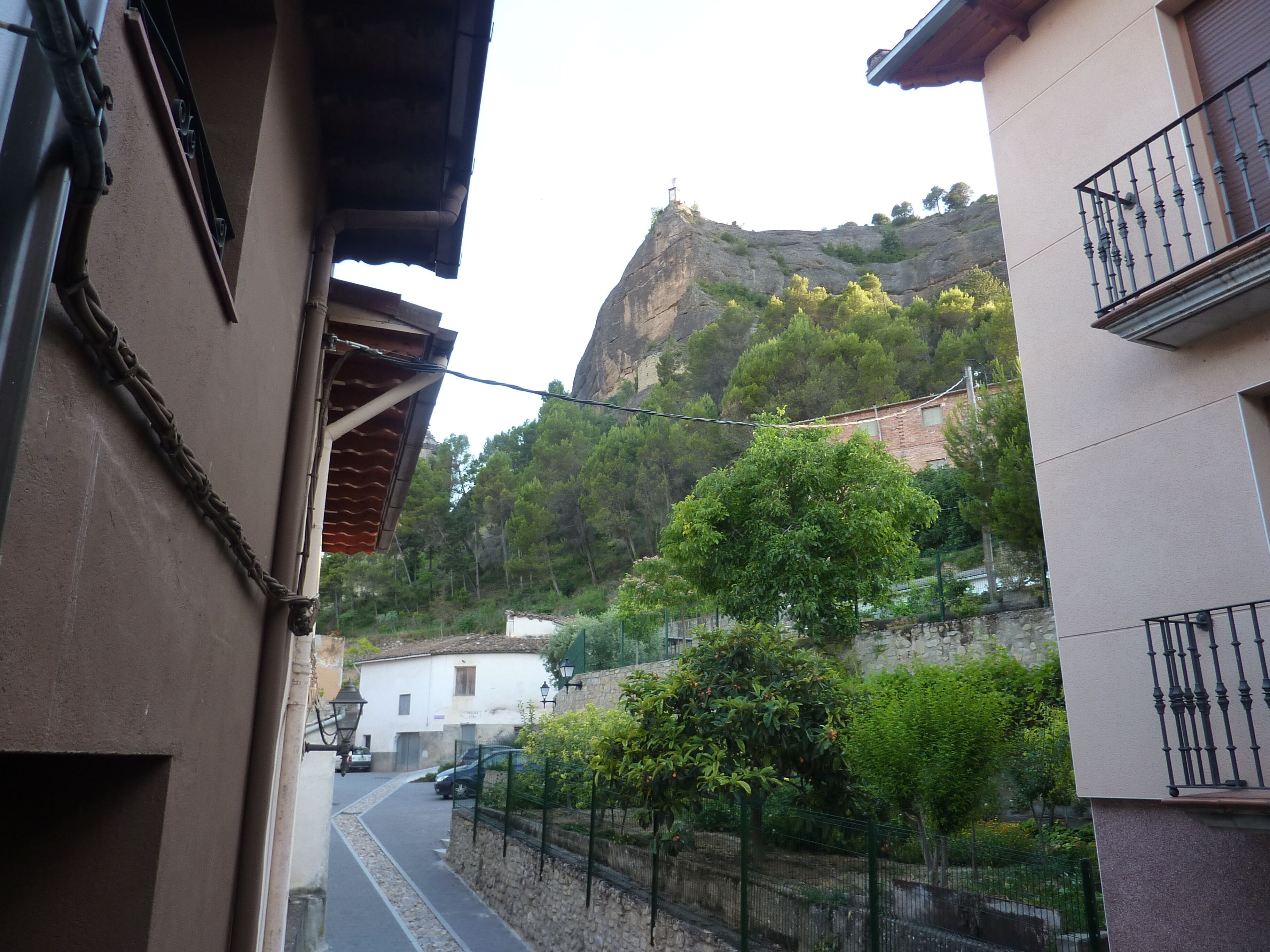 A narrow street with buildings on either side, a stone wall, and a view of a hill with trees.