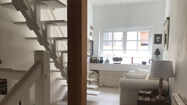 Una escalera conduce a una sala de estar con un sofá blanco, una lámpara y un ventanal con luz natural.