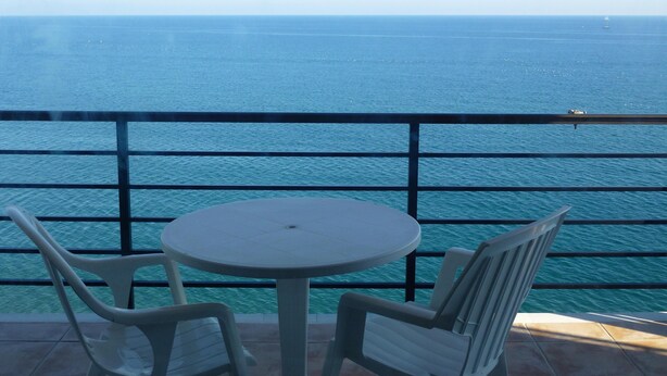 Ein Balkon mit rundem Tisch und zwei Stühlen, mit Blick auf ein ruhiges Meer und ein entferntes Segelboot.