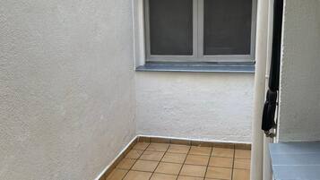 Un balcón pequeño y cerrado con suelo de baldosas, un estante metálico y una ventana con mosquitero.