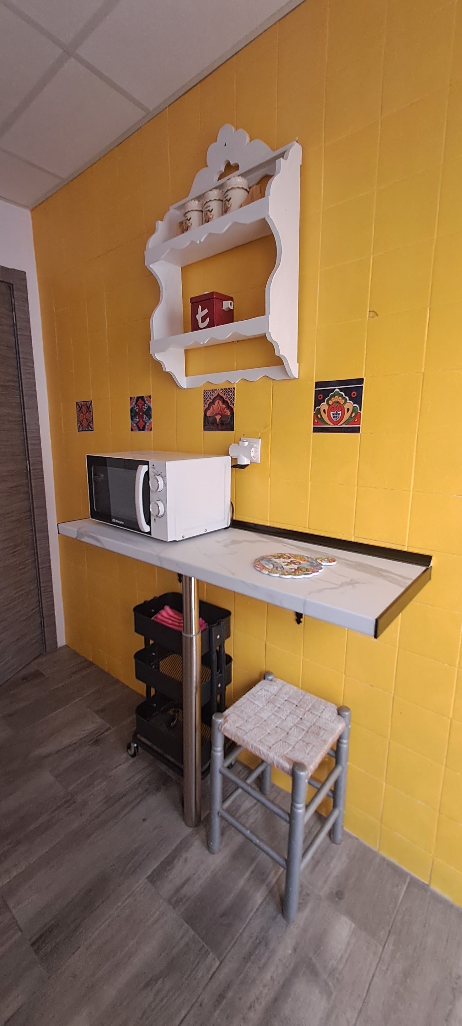 Une cuisine compacte équipée d’un four à micro-ondes, d’une petite table, d’un tabouret et d’une étagère où sont disposés des objets décoratifs.