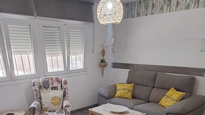 Un salon comprenant un fauteuil à motifs, un canapé gris avec des coussins jaunes, une table basse en bois et un luminaire suspendu.