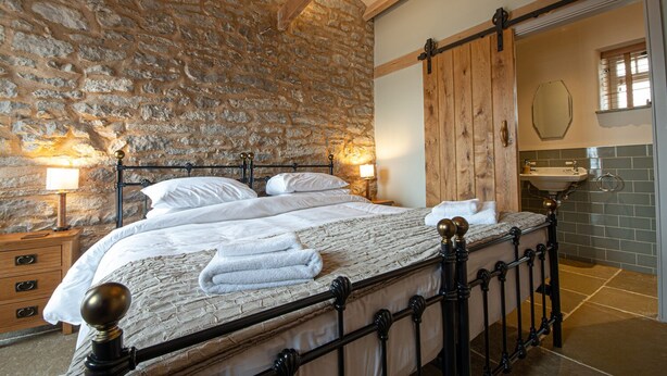 A bedroom with a stone wall, a wooden bed frame, a wooden door, and a bathroom with a sink and mirror.