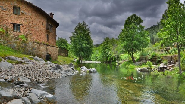 Un bâtiment en pierre, avec une rivière devant, entouré d’arbres et de verdure.