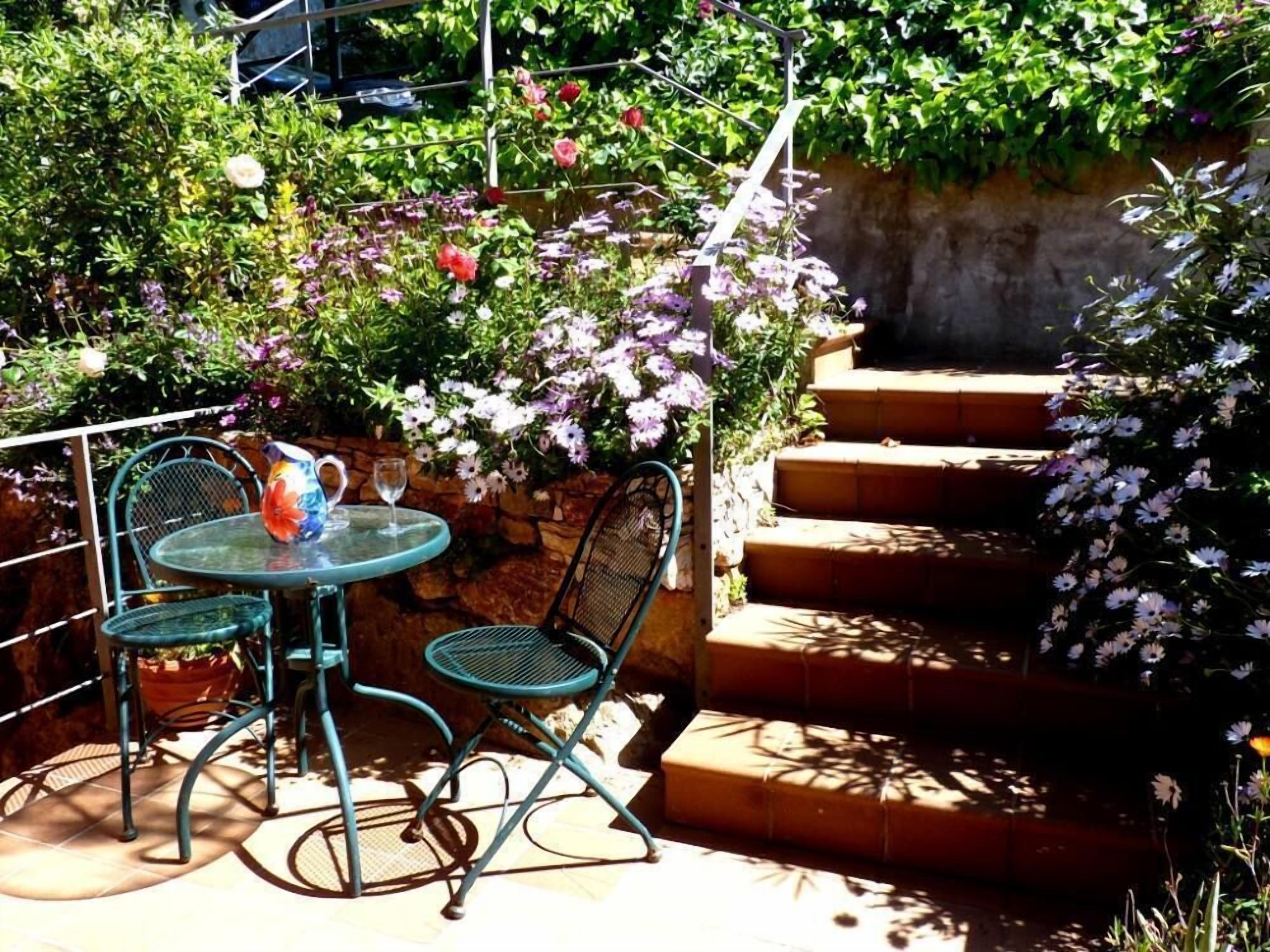 Un patio con una mesa redonda y dos sillas, rodeado de exuberante follaje y flores.