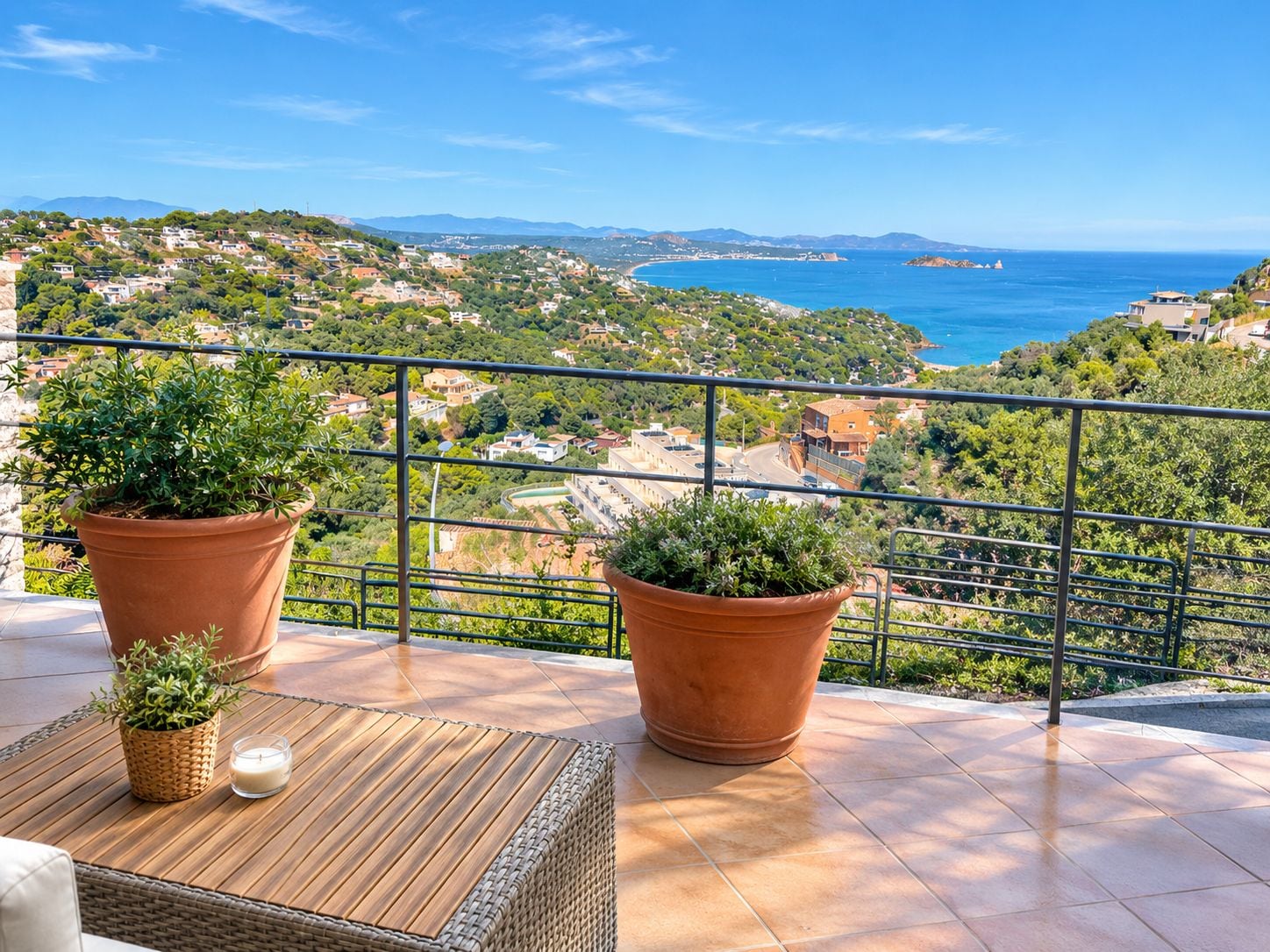 A balcony with terracotta pots, wicker furniture, and a view of a coastal town.