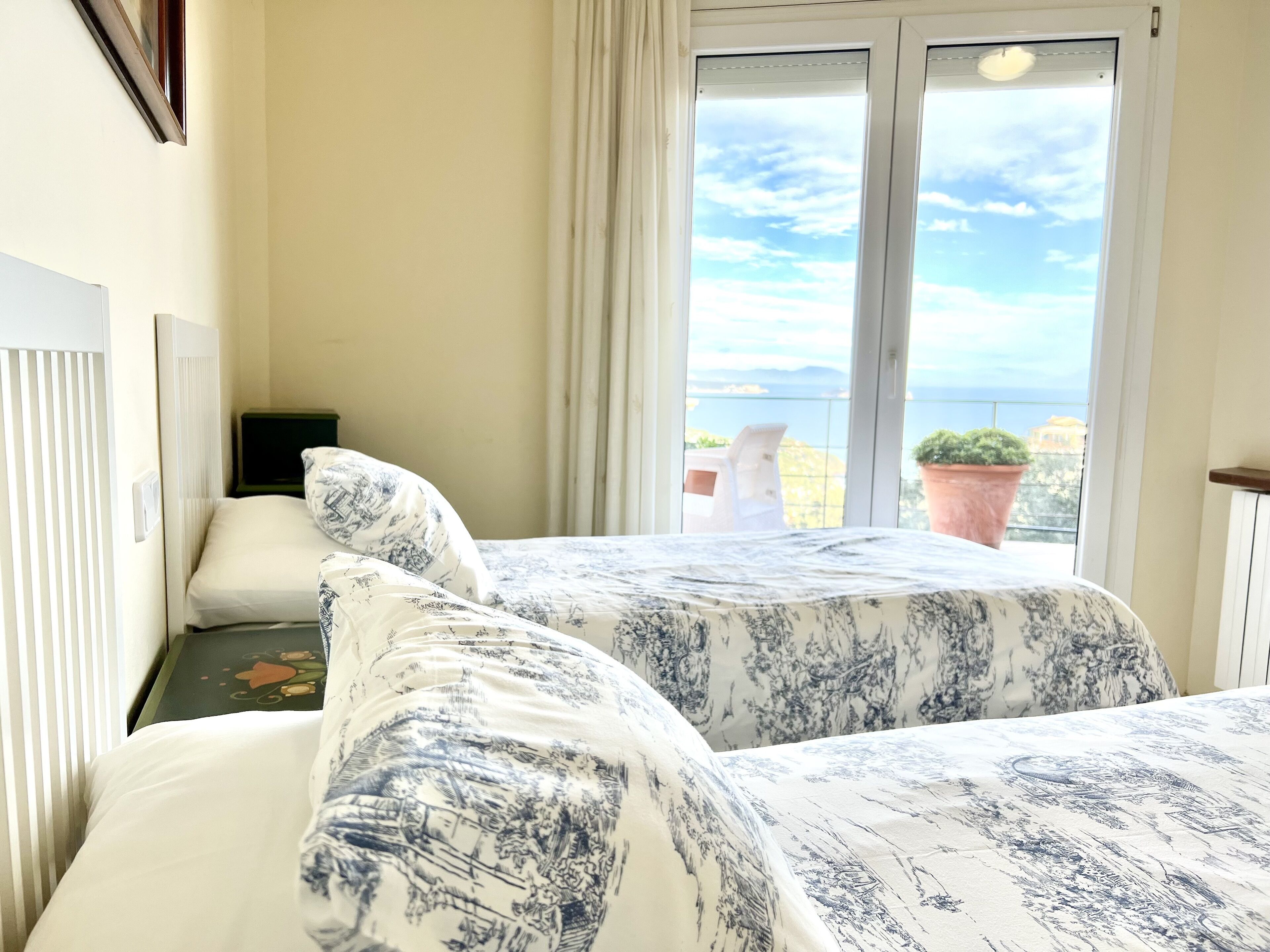 A hotel room with two beds, a window with a view of the sea, and a potted plant on the balcony.