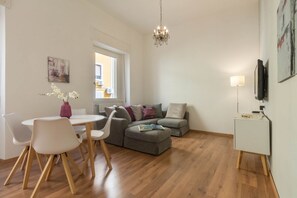 Een moderne woonkamer met een eethoek, een grijze zithoek, een kroonluchter en een flatscreen televisie aan de muur.
