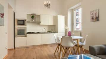 A modern kitchen with white cabinets, a microwave, and an oven. There is a dining area with a white table and chairs, and a book on a couch.