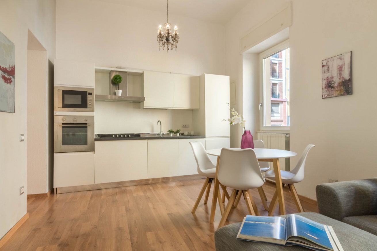 Eine moderne Küche mit weißen Schränken, einer Mikrowelle und einem Backofen. Es gibt eine Essecke mit einem weißen Tisch und Stühlen, und ein Buch auf einem Sofa.