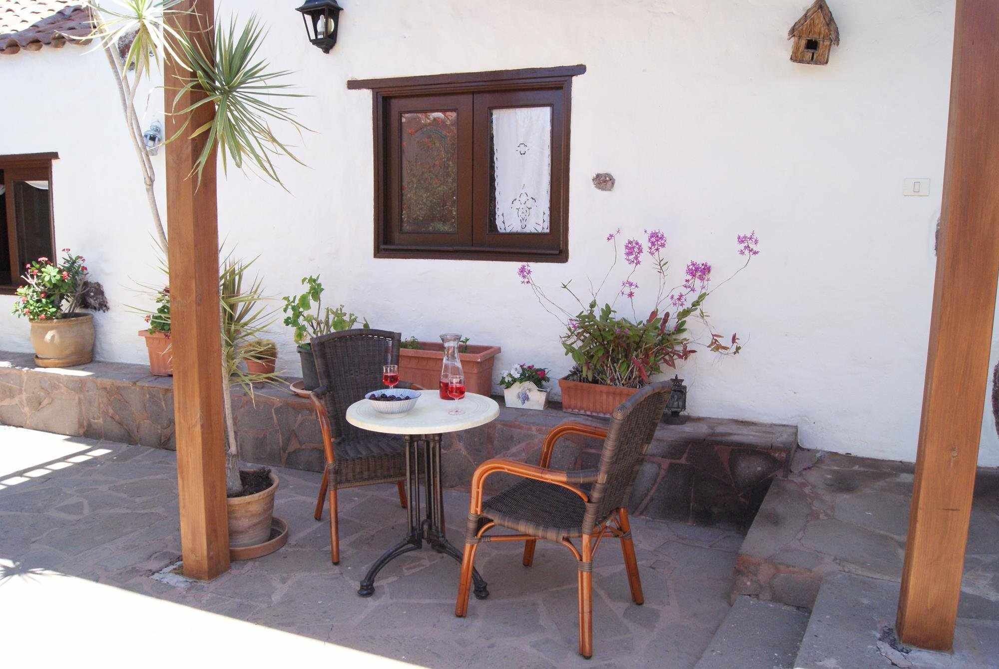 Un patio al aire libre con una mesa y dos sillas de mimbre, rodeado de plantas en macetas y una pequeña ventana con un marco decorativo.