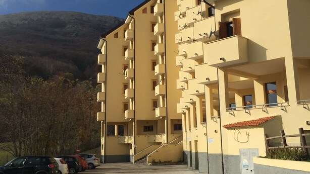 A multi-story building with balconies, a parking area, and a mountain in the background.