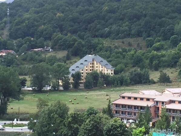 A large, multi-story building with a sloped roof, surrounded by lush greenery and hills.