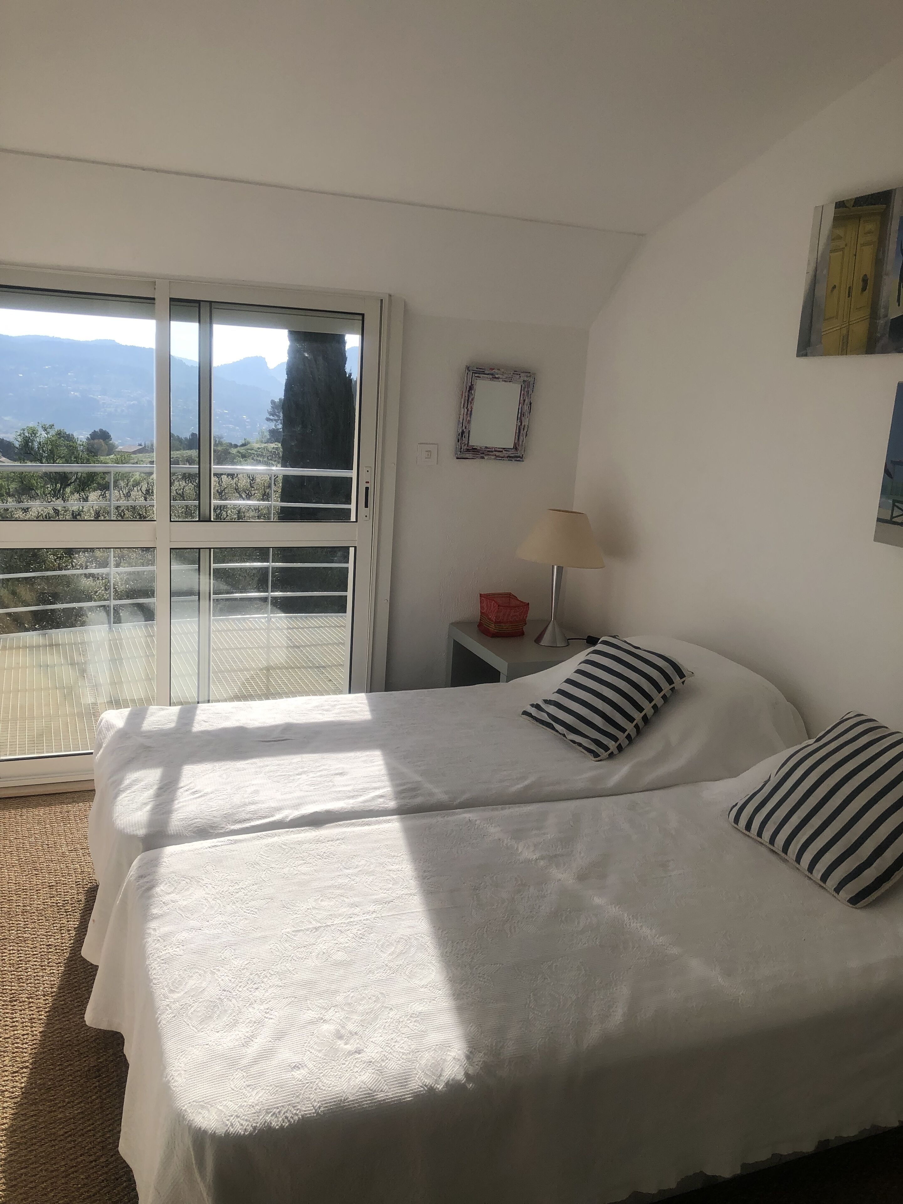 A bedroom with a bed, two pillows, a nightstand with a lamp, a framed mirror, and a view of mountains through a sliding glass door.