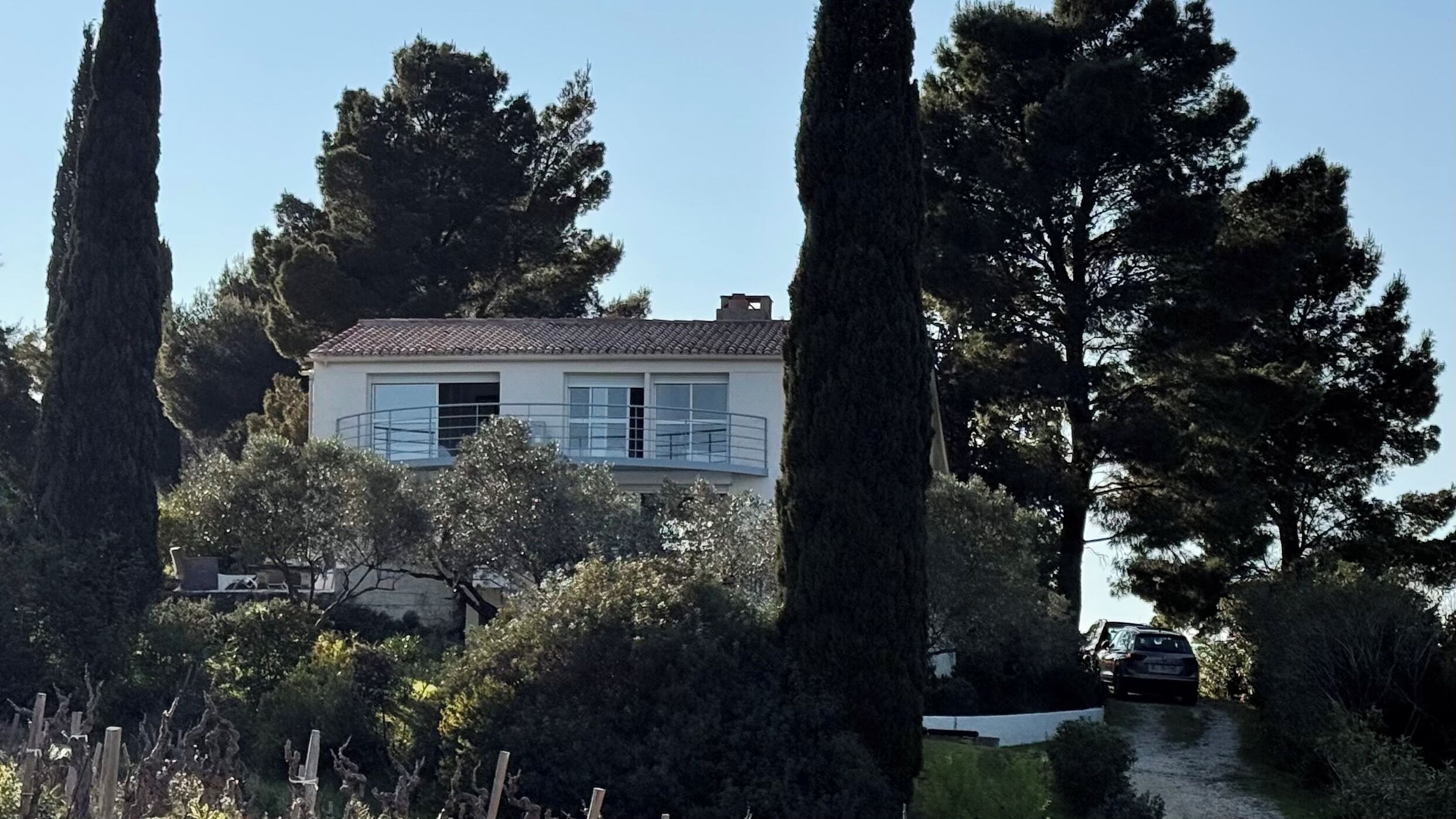A two-story house with a balcony, surrounded by tall trees and a gravel path.