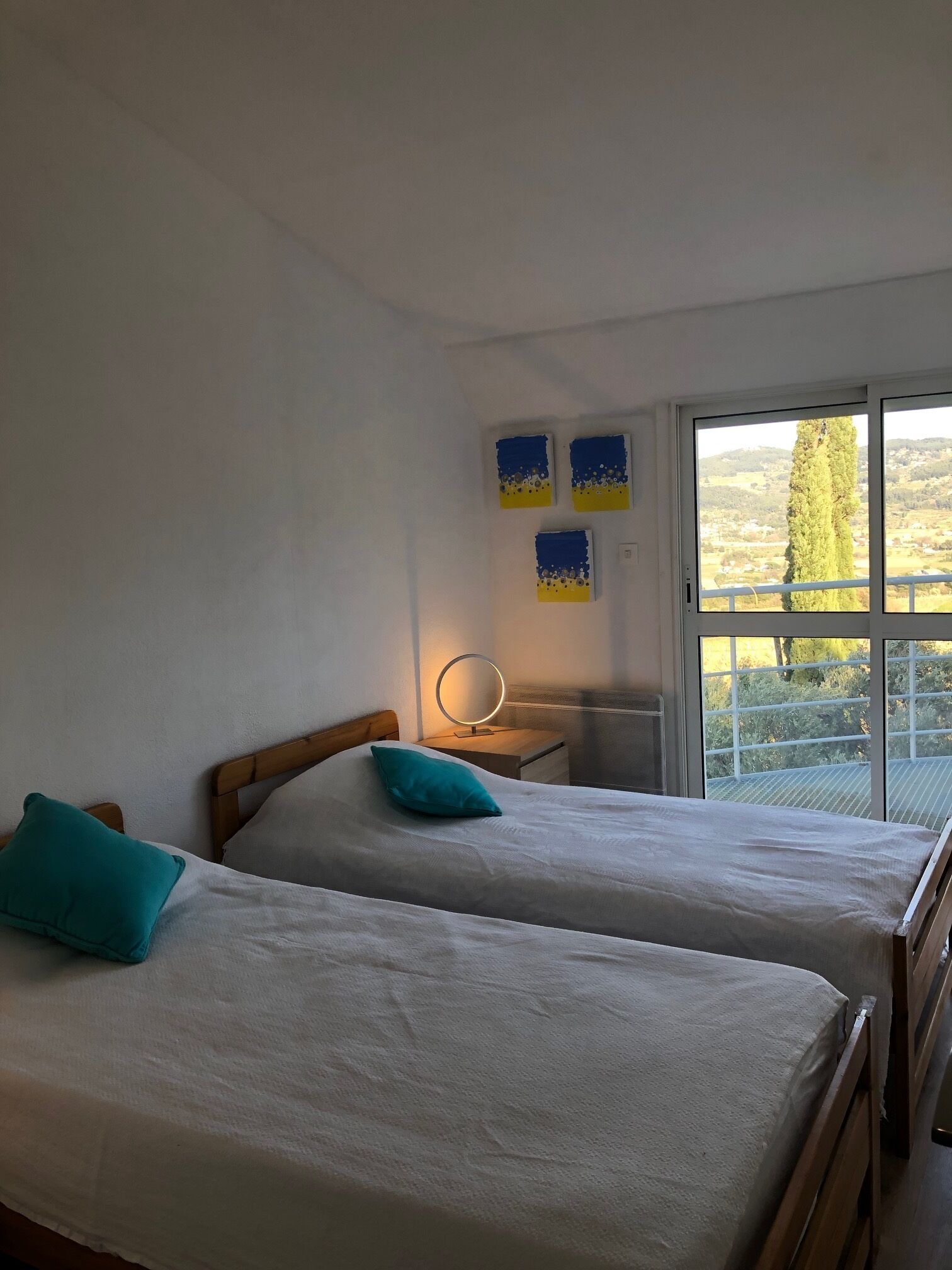 A bedroom with two beds, a bedside table with a mirror, and a window with a view of trees and hills.