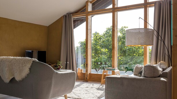 A modern living room with a sofa, a coffee table, and a large window with curtains. - StroEnzo - Cozy straw-bale holiday home - Tongeren / Maastricht - 5pers   (Riemst)