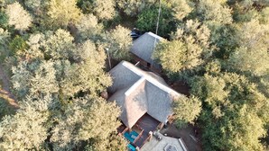 Vue aérienne d’une maison entourée d’une forêt dense, avec une grande terrasse et un jacuzzi.