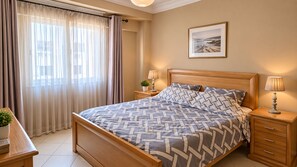 A neatly made bed with a patterned bedspread, a bedside table with a lamp, and a window with curtains.