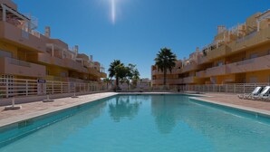 A swimming pool surrounded by multi-story buildings with balconies, palm trees, and a clear sky.