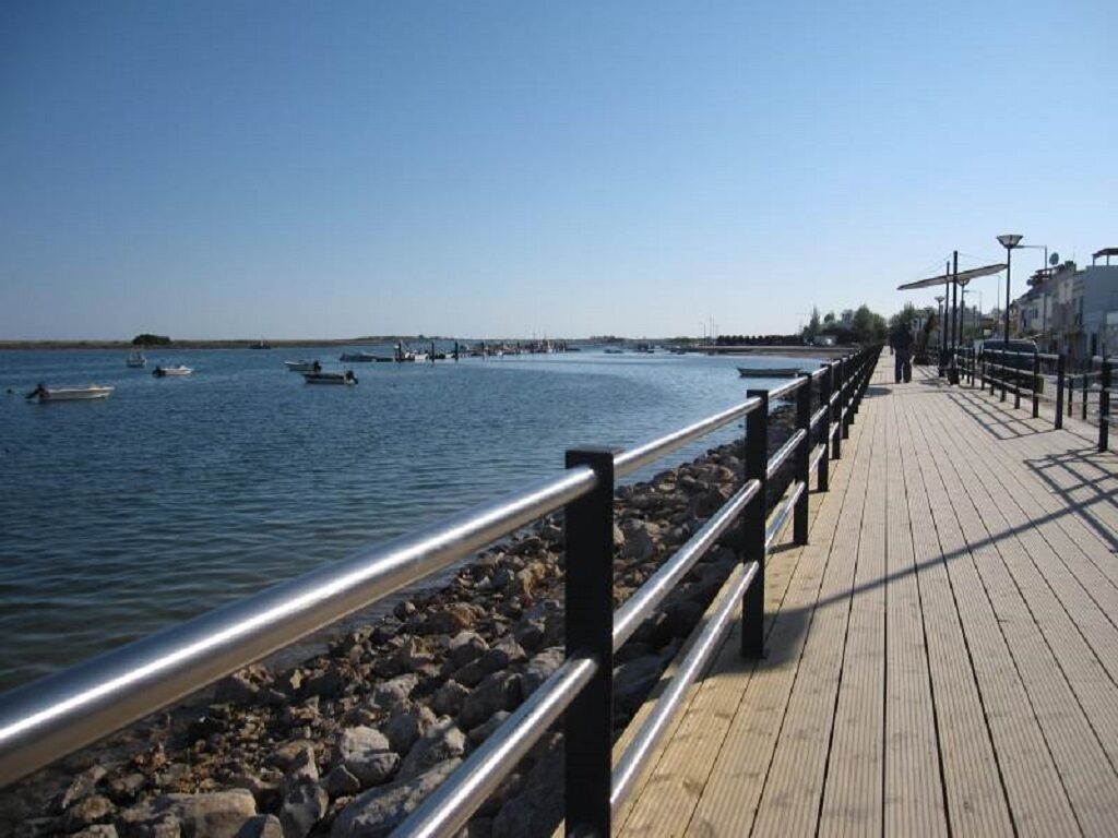 A wooden boardwalk by a calm body of water with boats and a clear sky.