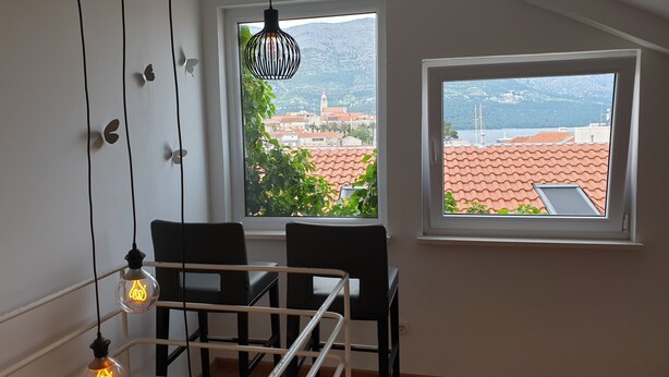 A room with a dining table and chairs, a window with a view of a cityscape, and hanging light fixtures. - Apartment Keti spacious apartment in the center of Korcula town with sea view (Korčula, Dubrovačko-neretvanska županija)