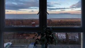 Vista desde una ventana hacia una playa, con una cerca y un coche aparcado.