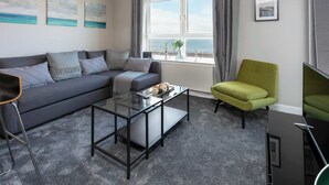 Un salón moderno con un sofá gris, una mesa de centro de vidrio y un sillón verde. Hay tres cuadros en la pared, una ventana con cortinas y vista al mar.