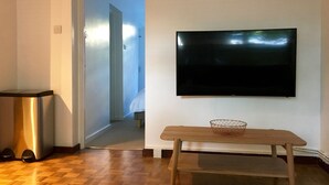 Una habitación con piso de madera, un televisor instalado en la pared, una mesita de madera con una cesta y un cesto de basura.