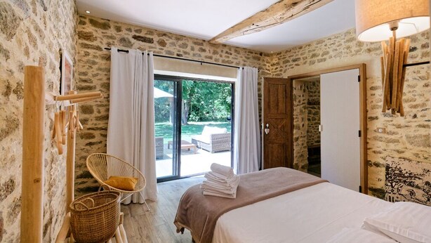 A bedroom with a stone wall, a bed with white linens, a wicker chair, and a view of an outdoor patio.