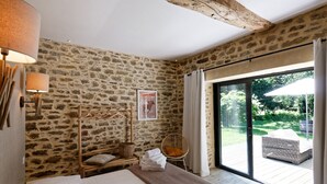 A bedroom with a stone wall, a bed with white linens, a wooden chair, and a view of the garden through a glass door.