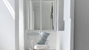 A white chair with a striped pillow in a corner of a room with white walls and a large window.