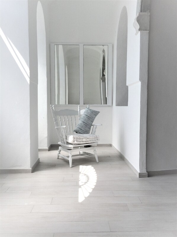 A white chair with a striped pillow in a corner of a room with white walls and a large window.