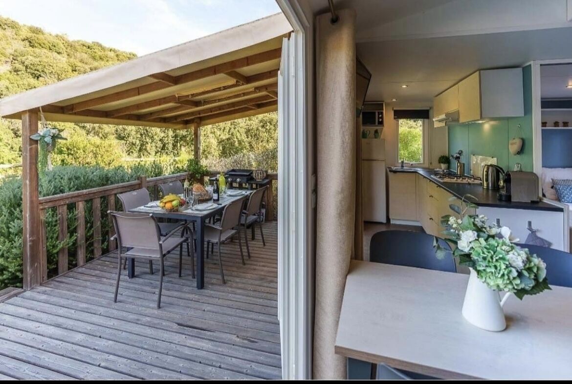 An outdoor dining area with a wooden deck, a covered seating area, and a kitchen visible through an open door.