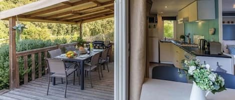 Un área de comedor al aire libre con terraza de madera, un espacio de estar techado y una cocina visible a través de un ventanal.